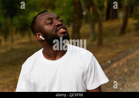 Glücklicher junger Erwachsener afroamerikanischer Mann, der mit Kopfhörern im Park spazieren geht Stockfoto