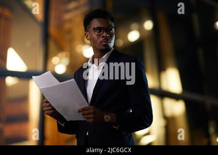 Seriös schöner afroamerikanischer Geschäftsmann mit Brille, der über die dunkle Straße läuft und die Papiere überprüft Stockfoto