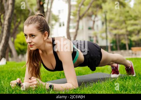 Junge, schöne Frau, die im Stadtpark sportliche Kleidung trägt, im Freien Plank-Übungen auf Yogamatte macht und sich nach vorne freut. Gesunder Lebensstil, Training Stockfoto