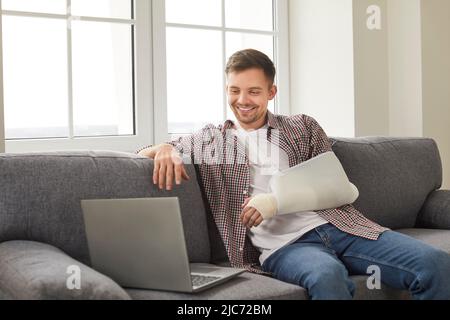 Mann mit gebrochenem Arm, der Filme auf einem Laptop ansieht oder zu Hause über einen Videolink mit Freunden spricht. Stockfoto