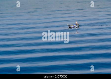 Aegina, Griechenland, - 2022. Mai: Person, die in einem kleinen Fischerboot auf einem blauen Meer mit Wellenmuster steht Stockfoto