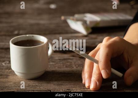 Eine Zigarette in den Händen eines Mädchens auf dem Tisch neben einer Tasse Kaffee, Zigaretten und Kaffee, schlechte Gewohnheiten, Rauchen Stockfoto