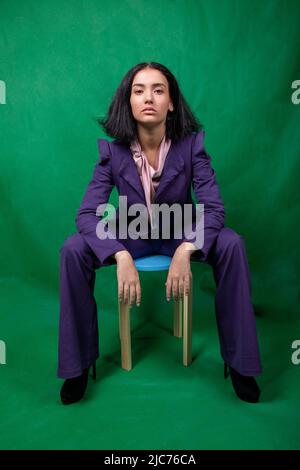 Junge hübsche Model sitzt auf einem blauen Hocker und ruht ihre Ellbogen auf den Knien im Studio Stockfoto