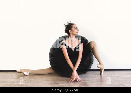 Anmutige Ballerina in schwarzem Schwanenkleid vor weißem Hintergrund. Junge Balletttänzerin, die vor der Aufführung in schwarzem Tutu, klassischem Tanzstud, praktiziert Stockfoto