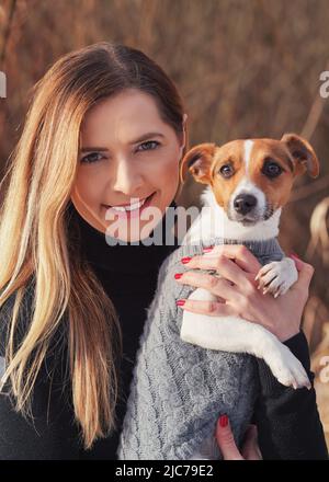 Junge Frau, die ihren Jack Russell Terrier Hund in warmer Winterkleidung an den Händen hält Stockfoto