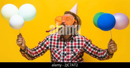 Bärtiger Mann im Geburtstagshut und lustige Brille mit Luftballons, die Noisemaker blasen. Partyzeit. Urlaub. Stockfoto