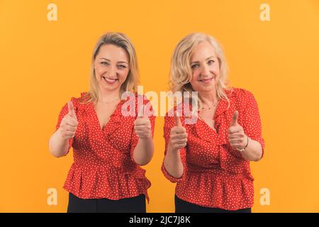 Das ist ausgezeichnet - zwei blonde kaukasische fröhliche Frauen zeigen Daumen nach oben orange Hintergrund Medium shot Studio Schuss Wohlbefinden Konzept. Hochwertige Fotos Stockfoto
