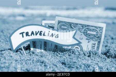 Papierrechnungen ein Dollar und kleine Stick Papier Sprechblase mit Wort Reisen begraben im Sand Strand Hintergrund Meer Nahaufnahme sonnigen Sommertag. Konzept Geld Reise Tourismus Urlaub. Blaue Farbe Stockfoto