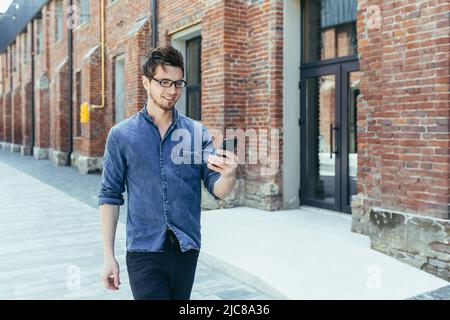 Ein junger, gutaussehender Mann mit Brille geht mit einem Mobiltelefon eine Stadtstraße entlang und sucht auf einem GPS-Navigator nach einem Platz und einer Straße. Stockfoto