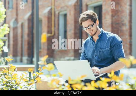 Junger Mann, Auszubildender. Er arbeitet online mit einem Projekt an einem Laptop und sitzt konzentriert auf einer Bank in der Nähe des Bürozentrums und trägt eine Brille. Stockfoto