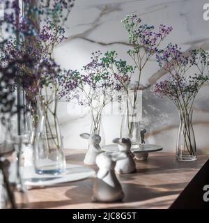 Blick auf die Einrichtung im modernen skandinavischen Stil. Mehrfarbige Gypsophila in Glasvasen und weißen Keramikhasen am Fenster. Osterkonzept. Wohnzimmer Stockfoto