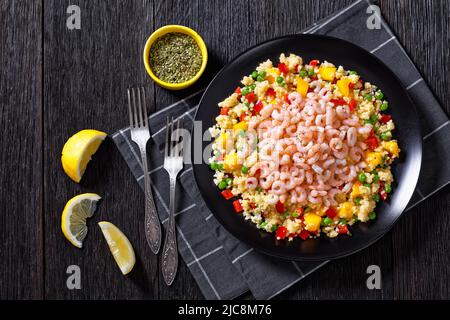 Gekochte kleine rosafarbene Garnelen auf Hirsepilaff mit Gemüse, Mangostückchen und Gewürzen auf schwarzem Teller auf dunklem Tisch, horizontale Ansicht von oben Stockfoto