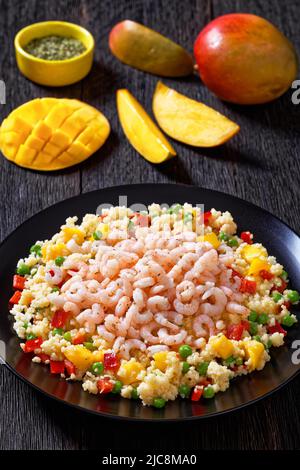 Gekochte kleine rosafarbene Garnelen auf Hirse-Pilaf mit Gemüse, Mangostückchen und Gewürzen auf schwarzem Teller auf dunklem Tisch mit Zutaten, Vertikal V Stockfoto