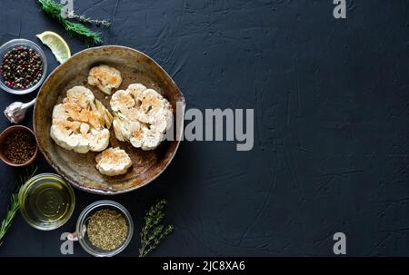 Blumenkohlsteak Kochen. Roher Blumenkohl, mit Gewürzen bestreut, liegt in einer Bratpfanne. Olivenöl, Kräuter, verschiedene Gewürze in der Nähe. Dunkler Hintergrund. Kopys Stockfoto