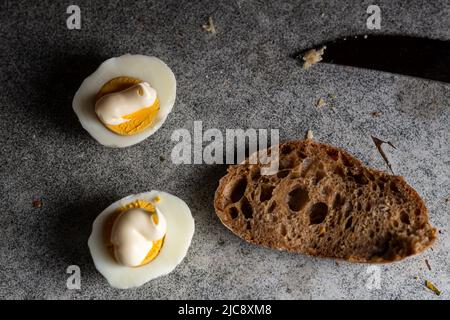 Hälften Hühnereier mit Mayonnaise und braunem Brot auf einem dunklen Küchentisch. Stockfoto