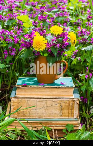 Braune Kaffeetasse mit einem Strauß Wildblumen auf einem Stapel alter Bücher auf einer Wiese mit blühenden Kräutern an einem sonnigen Morgen im Frühjahr oder Sommer aus nächster Nähe Stockfoto