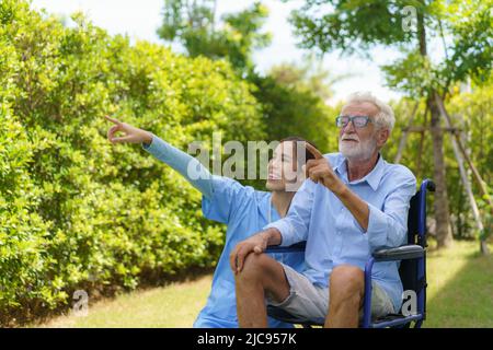 Asiatische Pflegekraft mit einem älteren Mann, der morgens im Garten auf dem Rollstuhl sitzt und die Natur nach Hause schaut. Stockfoto