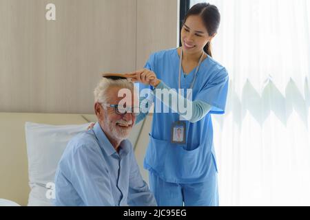 Junge asiatische Krankenschwester hilft, Haare zu putzen behinderten älteren Mann im Bett im Altersheim. Tausendjährige Pflegekraft, die behinderten Senioren hilft Stockfoto