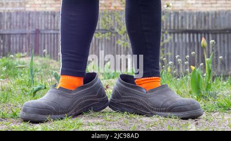 Frauenbeine in übergroßen Gummigaloshes vor dem Hintergrund der Landschaft. Beine in schwarzen Leggings, orangefarbenen Socken und schmutzigen Galoschen in einem Vorort Stockfoto