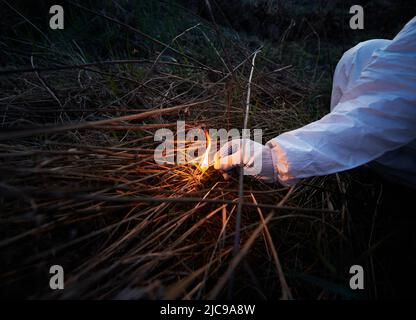 Mann, der altes getrocknetes Gras auf dem Feld verbrennt. Nahaufnahme der Hand, die brennendes Streichholz hält und nachts trockenes Gras in Brand setzt. Konzept der Ökologie und des menschlichen Faktors in Bränden. Stockfoto