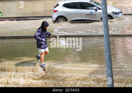 Junger Mann, der versucht, auf einer überfluteten Straße mit Autos auf dem Hintergrund zu gehen. Schmutziges Wasser nach starkem Regen Stockfoto