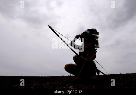 Ein junges indianisches Mädchen aus der Urzeit wird vor dem Abendhimmel silhouettiert. Sie steht stolz mit ihren Waffen an ihrer Seite. Stockfoto