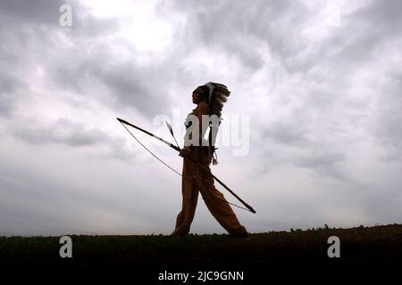 Ein junges indianisches Mädchen aus der Urzeit wird vor dem Abendhimmel silhouettiert. Sie steht stolz mit ihren Waffen an ihrer Seite. Stockfoto