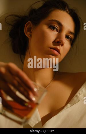 Junge Frau trinkt Alkohol aus Glas vor sonnigem Wandhintergrund, sucht Stockfoto