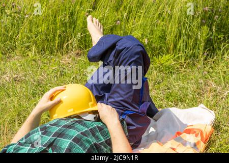 Arbeiter entspannt sich auf einer Wiese mit Helm und in Arbeitskleidung, Konzept, Work Life Balance, ausgewogene Arbeits- und Ruhezeiten in verschiedenen Beruf Stockfoto