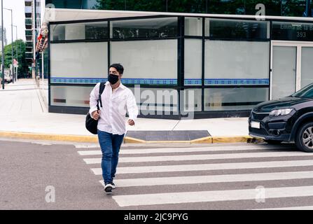 Junger hispanischer Mann mit Maske für Covid-19 beim Überqueren der Straße. Stockfoto