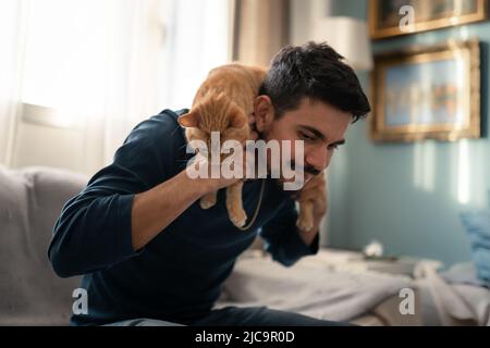 Der junge Mann spielt auf einem Sofa mit einer braunen gestromten Katze Stockfoto