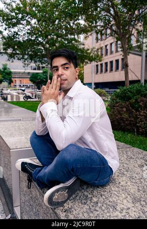 Attraktiver lateinischer Mann in einem weißen Hemd, der auf einem Granit mit einem Baum im Hintergrund sitzt. Stockfoto