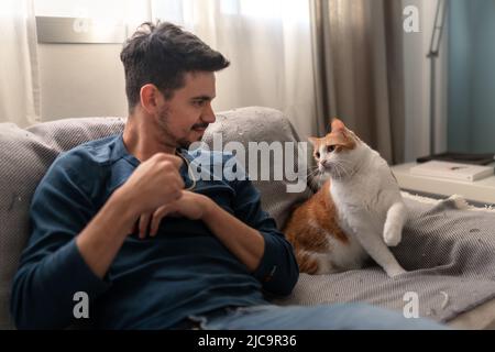 Der junge Mann spielt auf einem Sofa mit einer braunen und weißen Katze Stockfoto