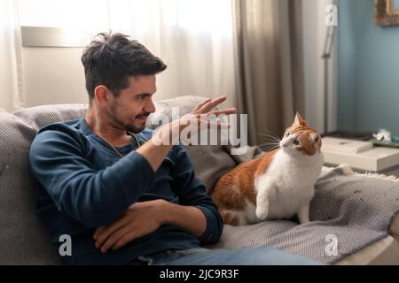 Der junge Mann spielt auf einem Sofa mit einer braunen und weißen Katze Stockfoto