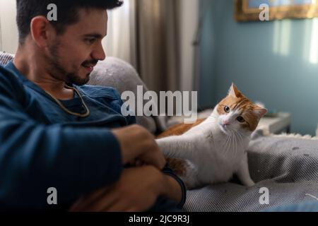 Der junge Mann spielt auf einem Sofa mit einer braunen und weißen Katze Stockfoto