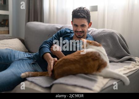 Der junge Mann spielt auf einem Sofa mit einer braunen und weißen Katze Stockfoto