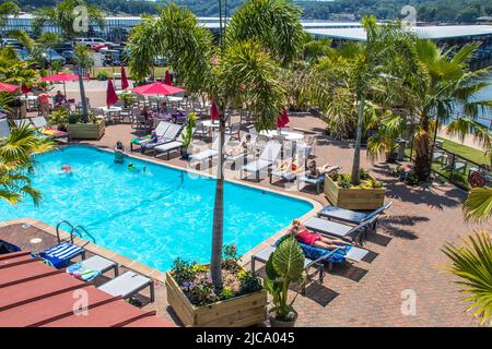 2021 - 06 -20 Grand Lake, OK USA - tropische Bäume um den Swimmingpool mit Frauen-Sonnenbaden und Bootsanlegestellen und geparkten Autos in der Ferne - Strandtuch Stockfoto