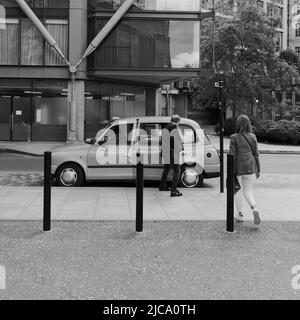 London, Großraum London, England, Juni 08 2022: Monochrom. Ein Mann öffnet eine Taxitür, als er und eine Begleiterin kurz vor dem Eingang stehen. Stockfoto