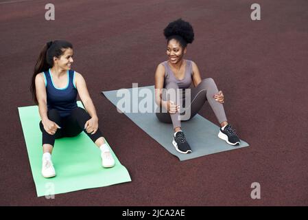 Trainingspause. Zwei unterschiedliche junge Frauen sitzen auf Yogamatten im Stadion und reden lachend. Stockfoto