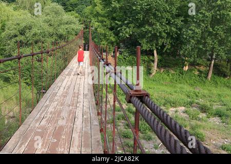 Eine alte hölzerne lange Hängebrücke über den Belaya-Gebirgsfluss, ein Junge in einem roten T-Shirt geht entlang der Brücke. Sonniger Sommertag Stockfoto