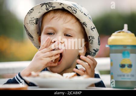 Nahaufnahme Bild des kaukasischen Jungen mit blonden Haaren und Hut Öffnung Mund gehen knusprig Brot beißen, mit freudigen Gesichtsausdruck. Kindheit, fo Stockfoto