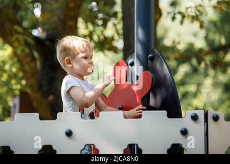 Der mutige kleine Junge spielt den Kapitän des Schiffes, er dreht das rote Ruder des Schiffes. Moderner, moderner Spielplatz in einem grünen Sommer-Eichenpark. Stockfoto