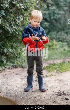 Ein kleiner Junge erkundet den Schlamm in der Nähe einer Pfütze während eines Spaziergangs in der Natur, er schaut auf seine verschmierten Hände. Stockfoto