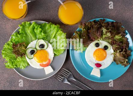 Fun Food für Kinder - lächelnde Gesichter aus Spiegeleiern, Gurken, Tomaten, Radieschen, schwarzen Oliven und Salatblättern auf zwei Tellern. Kreativ gesund Stockfoto