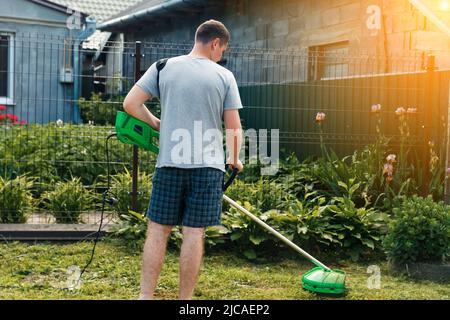 Rasentrimmer. Rückansicht eines Mannes, der das Gras mäht. Außenansicht eines jungen Arbeiters mit einem Rasenmäher, der Gras in einem unscharfen Hintergrund schneidet Stockfoto