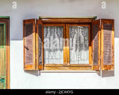 Altes Holzfenster mit offenen Fensterläden an weißer Stuckwand mit aufgehauenen Insektengeflechten und Pfauenvorhängen aus weißer Spitze - Teil der Tür sichtbar - golde Stockfoto