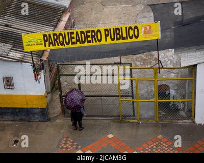 Medellin, Antioquia, Kolumbien - 6 2022. März: Eine Frau mit einem schwarzen Regenschirm und Fuchsia-Flecken wartet am Tor unter einem gelben Schild mit der Aufschrift „Pub“ Stockfoto