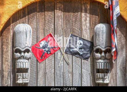 Zwei rustikale Holzskelettköpfe aus Tiki-gott hängen an einer rauen Holzwand in einem Stuckbogen mit Piratenfahnen, die umragen - Horror-Piratenhintergrund Stockfoto