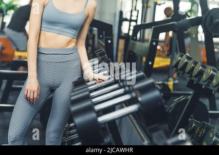 Nicht erkennbare kaukasische schlanke Frau, die Sport-BH und gemusterte Leggings trägt und nach einer großen Hantel greift. Innenaufnahme. Hochwertige Fotos Stockfoto