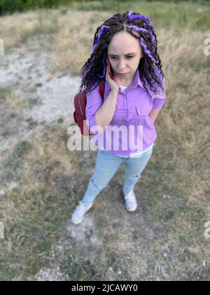 Junge schöne Frau, die auf dem Handy spricht und in hügeligem Gelände steht. Niedliche lebendige weibliche mit Dreadlocks sprechen auf dem Smartphone, im Freien stehen Stockfoto